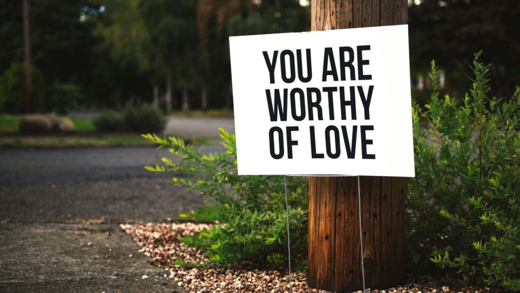 Eifersucht und Selbstliebe: Ein Schild steht am Straßenrand vor einem Baum. Auf dem Schild steht: You are worthy of love