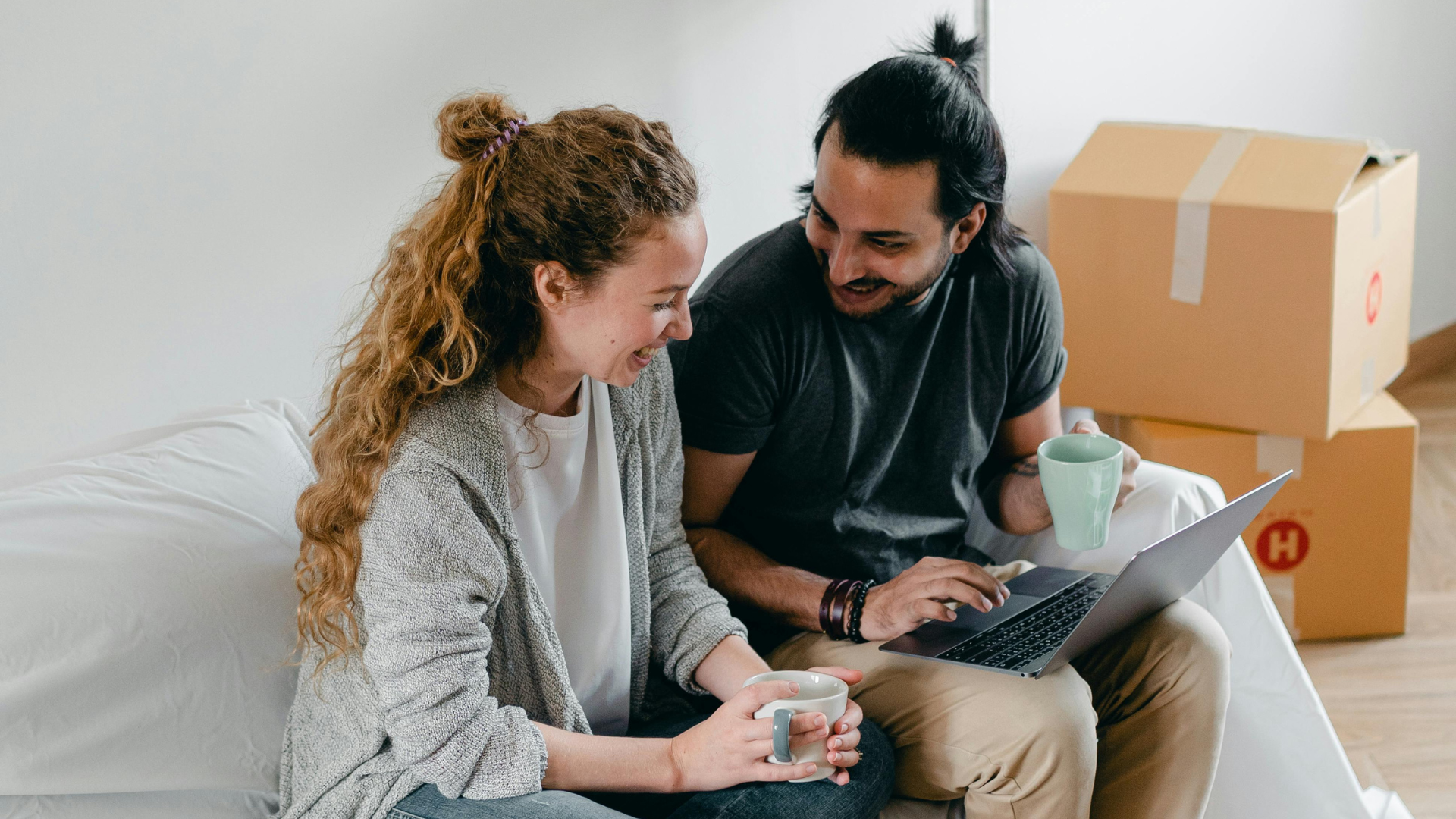 Zukunftsinvestment: Eine Frau und ein Mann sitzen lächelnd auf einer Matratze nebeneinander, er hat einen Laptop am Schoß, daneben sind Umzugskartons.