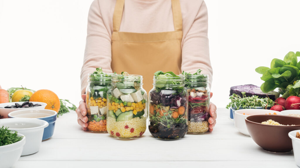 Eine Frau hält 4 Mahlzeiten in Schraubgläsern: Salate, Bohnen, Quinoa, Mais, Tofu, Schafskäse – perfektes Meal Prep