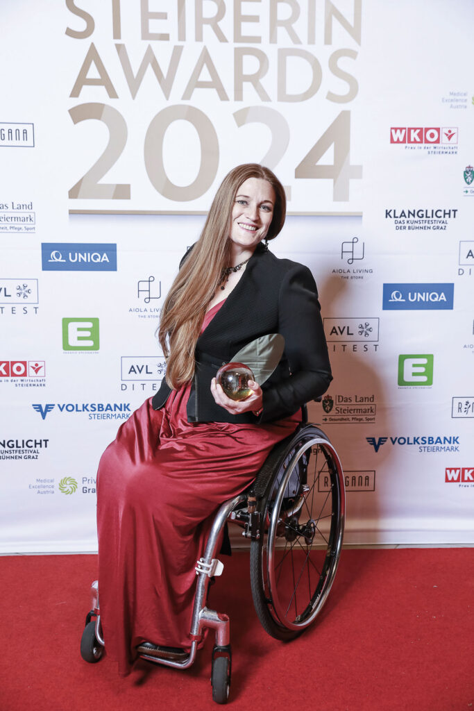 Barbara Sima-Ruml sitzt in einem eleganten, roten Kleid im Rollstuhl und hält den STEIRERIN AWARD in der Hand.