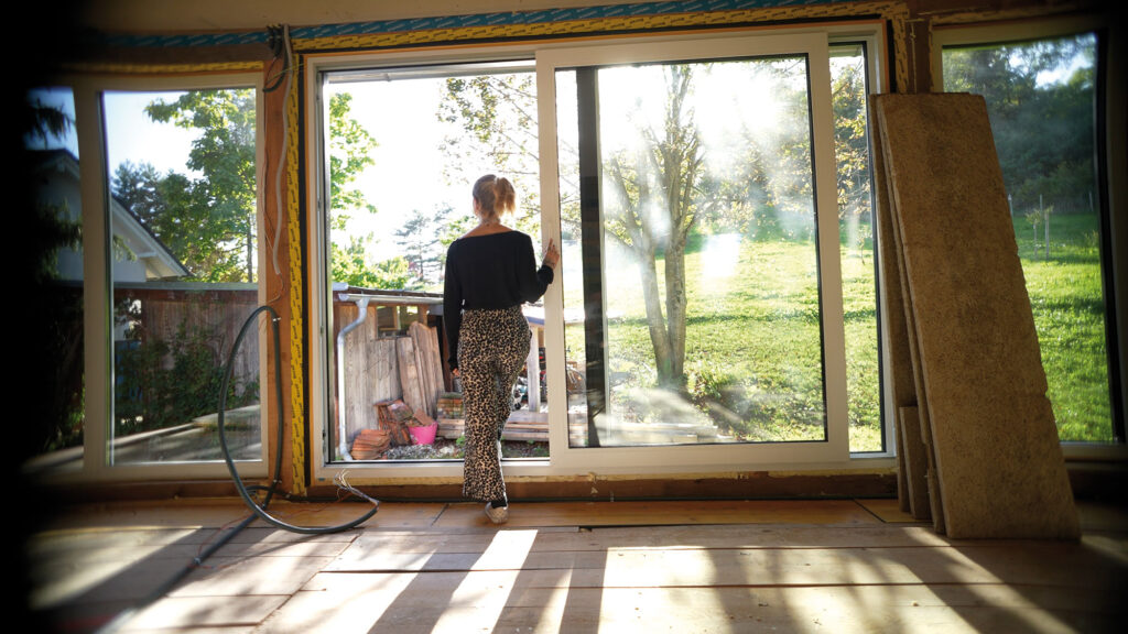 Frau von hinten steht in leerem Raum vor großer Fensterfront und sonnigem Garten.