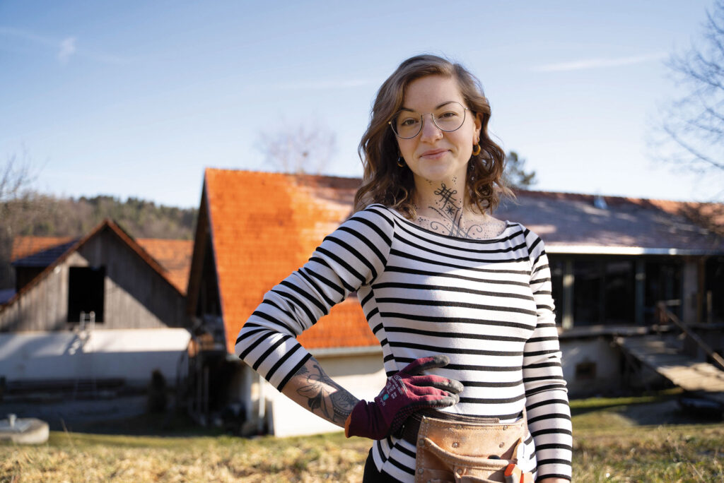 Portrait von Lisa M., „Die Hauswerkerin“ im Freien vor einem Haus mit gestreiftem Shirt.