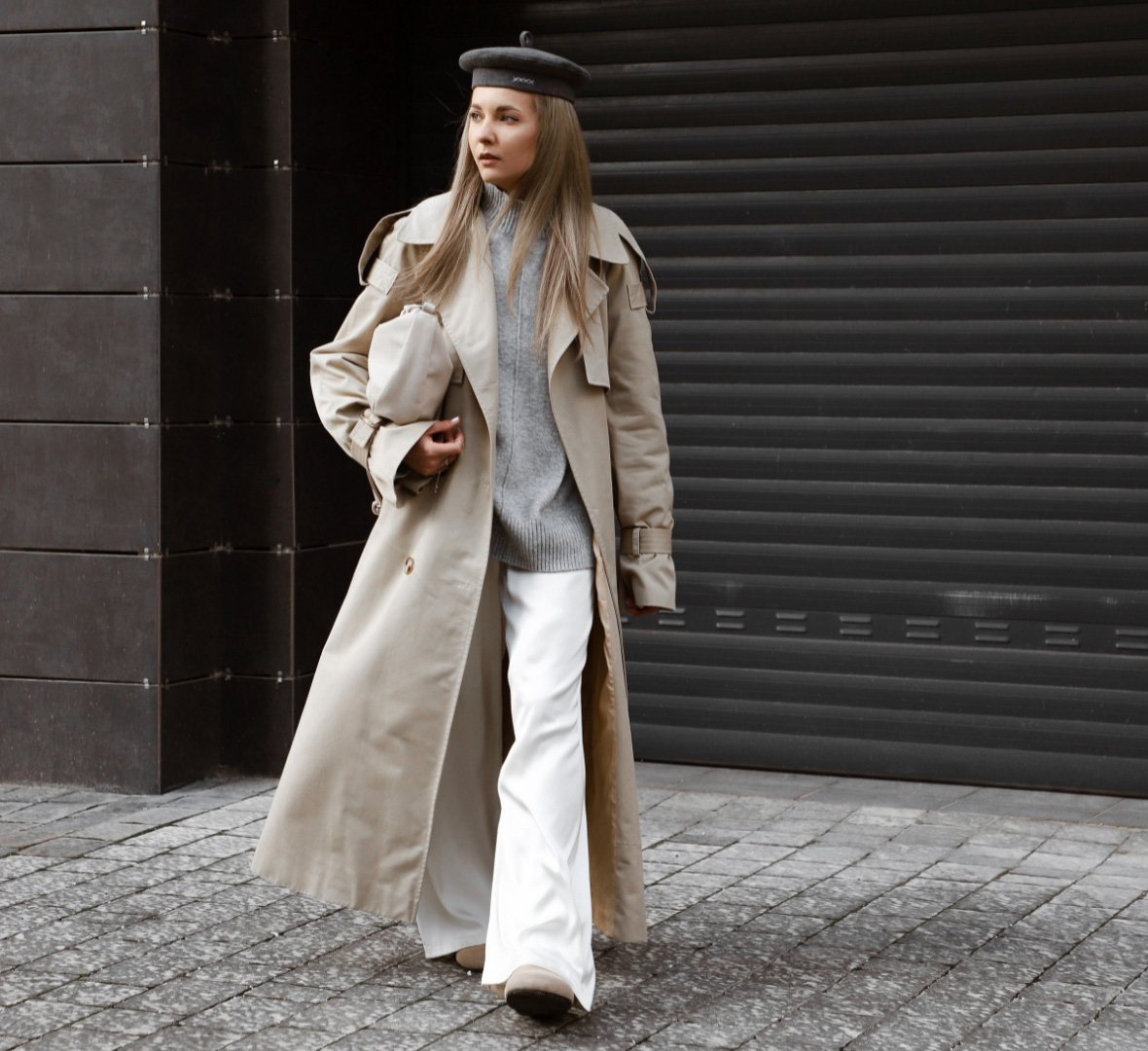Satinhosen Trend: Eine blonde Frau trägt eine Satinhose, einen Trenchcoat, grauen Pulli und Baskenmütze. Sie geht auf die Kamera zu