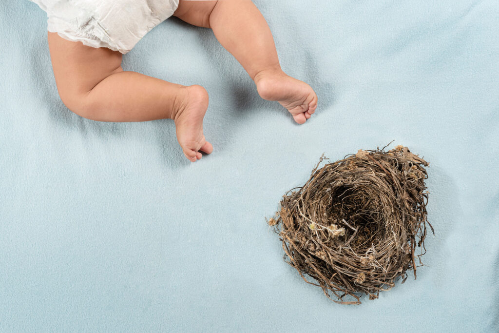 Loslassen: Beine von einem kleinen Baby mit Windel auf blauer Decke und Vogelnest daneben.