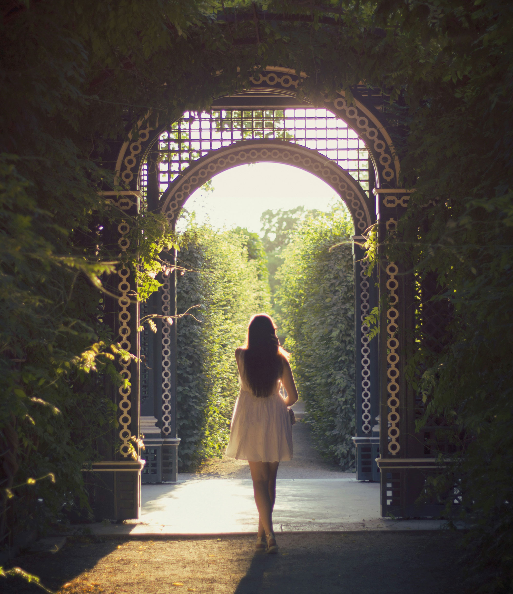 Frau geht in leichtem Sommerkleid barfuß durch ein großes Tor in ein Gartenlabyrinth.