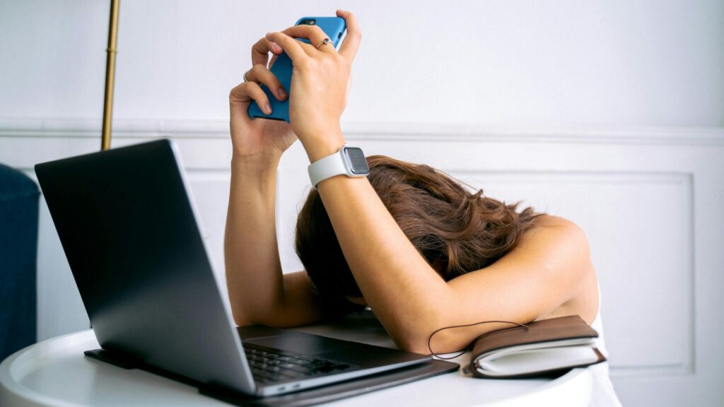 Digitale Balance: Frau hat beide Ellenbogen auf einen Tisch gestützt, ihr Kopf liegt nach unten gerichtet auf dem Tisch. Vor ihr steht ein Laptop. in ihren Händen hält sie ein Handy.