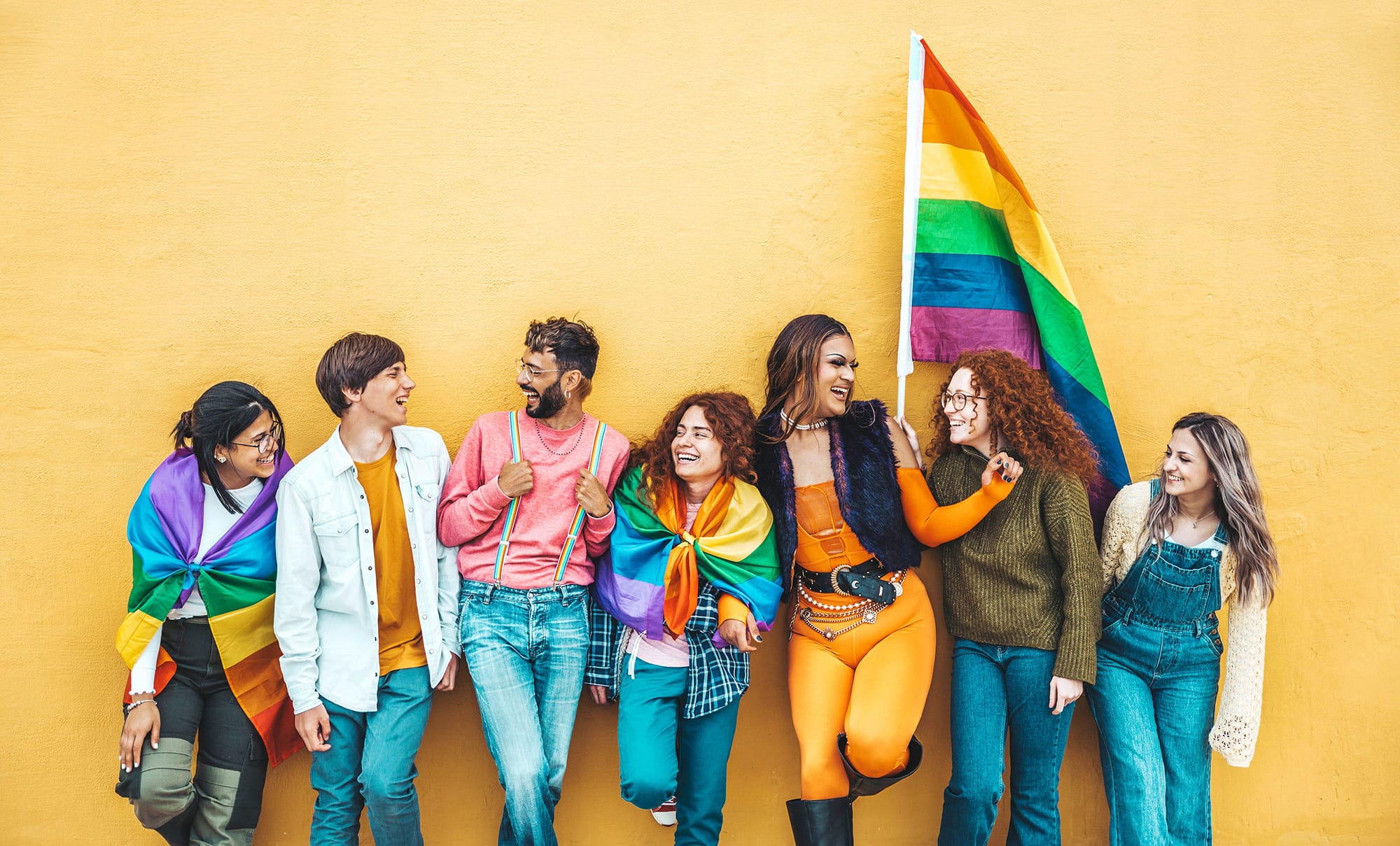 Eine Gruppe queerer Menschen steht lachend for einer gelben Wand mit einer großen Regenbogenflagge in der Hand.