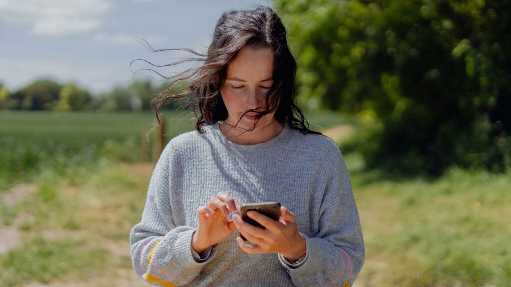 Kinkeeping: Frau steht in der Natur und tippt mit gesenktem Blick auf ihr Handy.