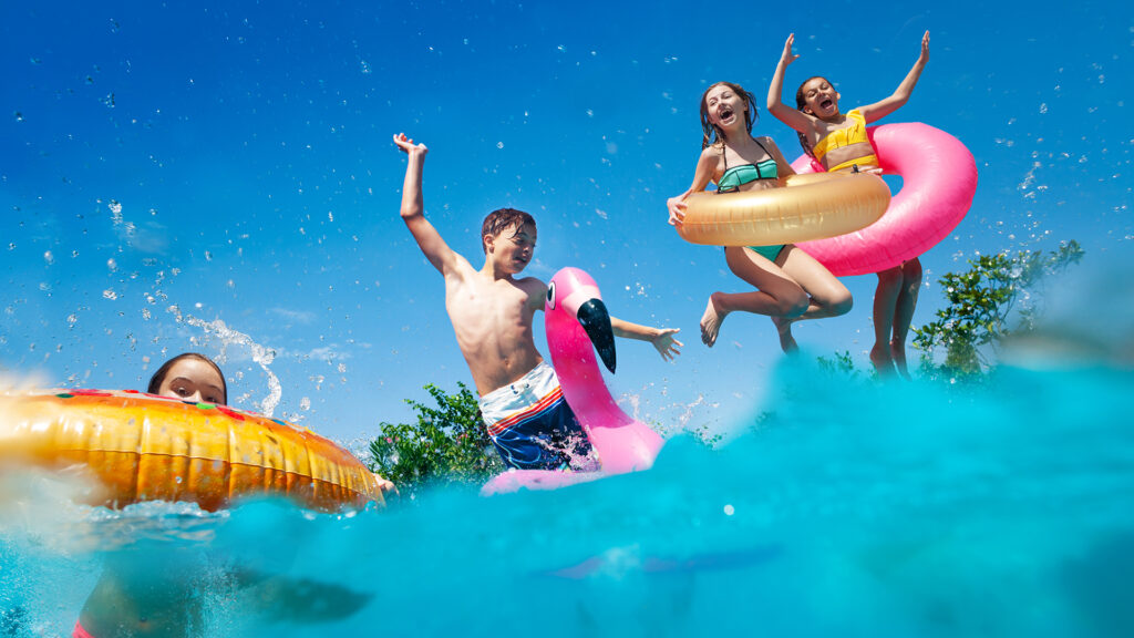 Halb unter Wasser aufgenommenes Bild von vielen kleinen Kindern, die im Schwimmbecken tauchen, aufblasbares Spielzeug werfen, die Hände heben und Spaß haben.