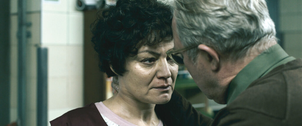Filmszene mit älterem Mann, der einer Frau von hinten an Hals fasst und ihr dabei intensiv in die Augen schaut.