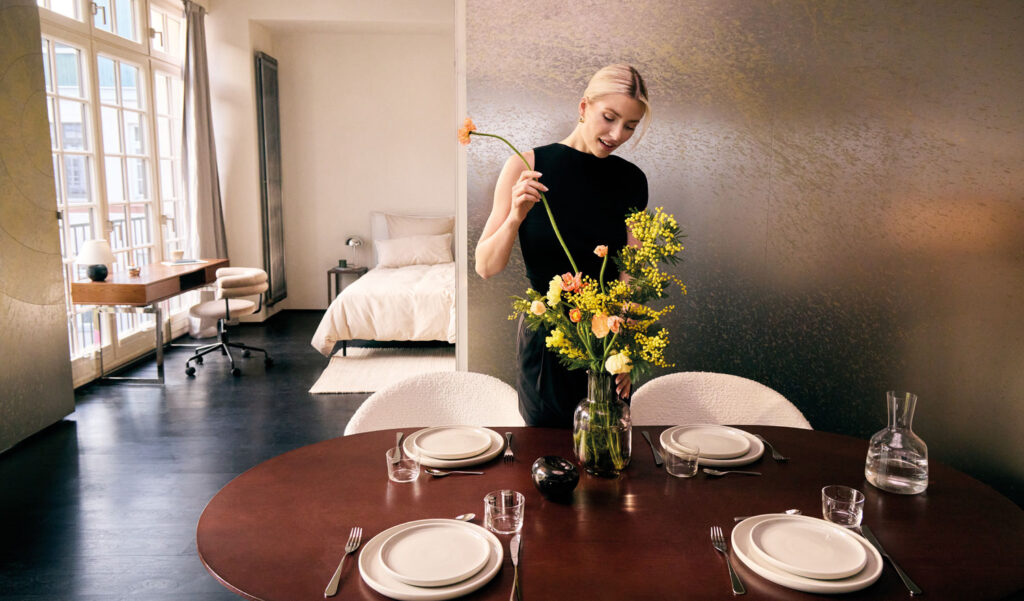 Individuelles Wohndesign: Frau steht gelbe Blumen in Vase auf einem Esstisch in einem großen Raum mitt Bett und Schreibtisch.