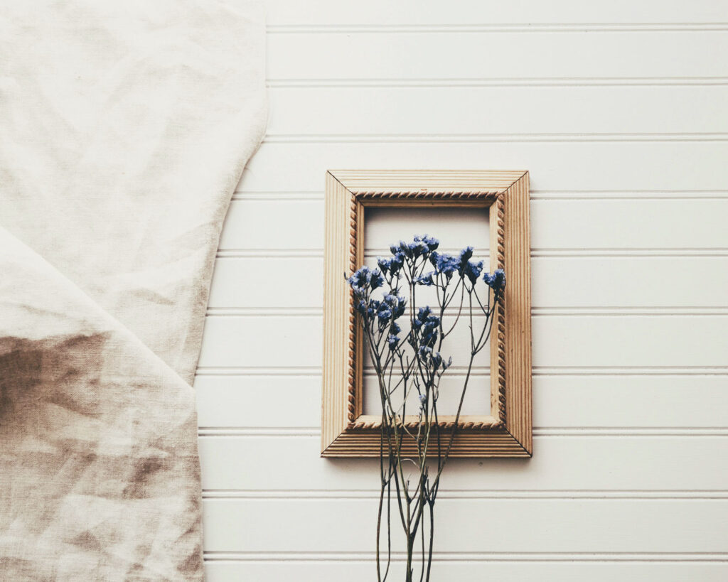 Hölzerner Bilderrahmen mit blauen getrockneten Blumen davor auf heller Täfelungs-Wand.