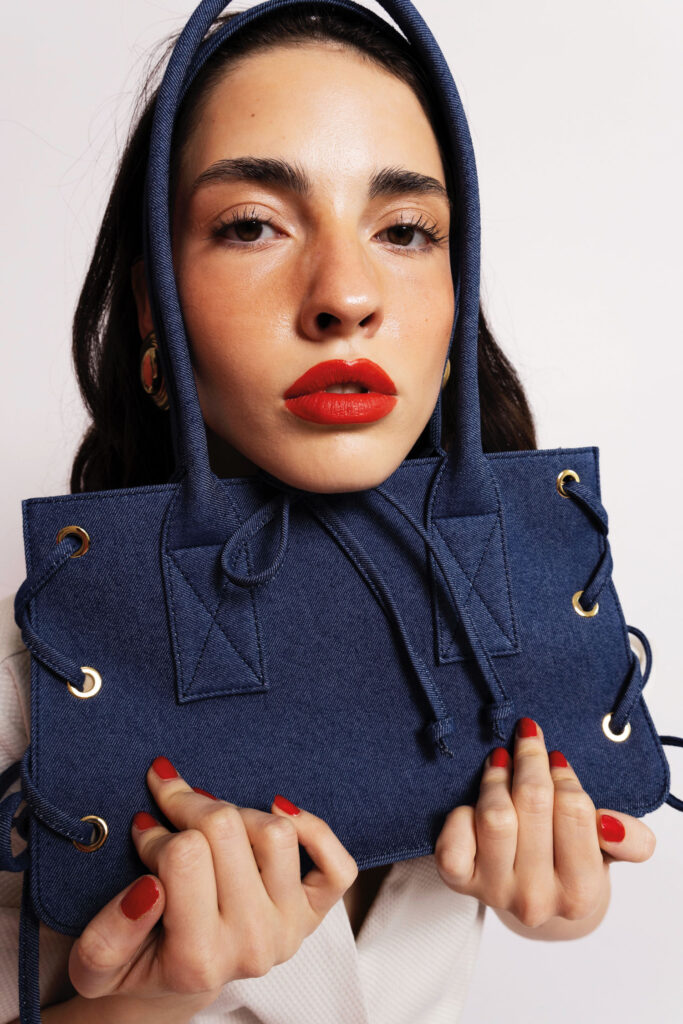 Julia Skergeth Handtaschen: Blaues Jeansmodell präsentiert von einer dunkelhaarigen Frau mit roten Lippen und Nägeln