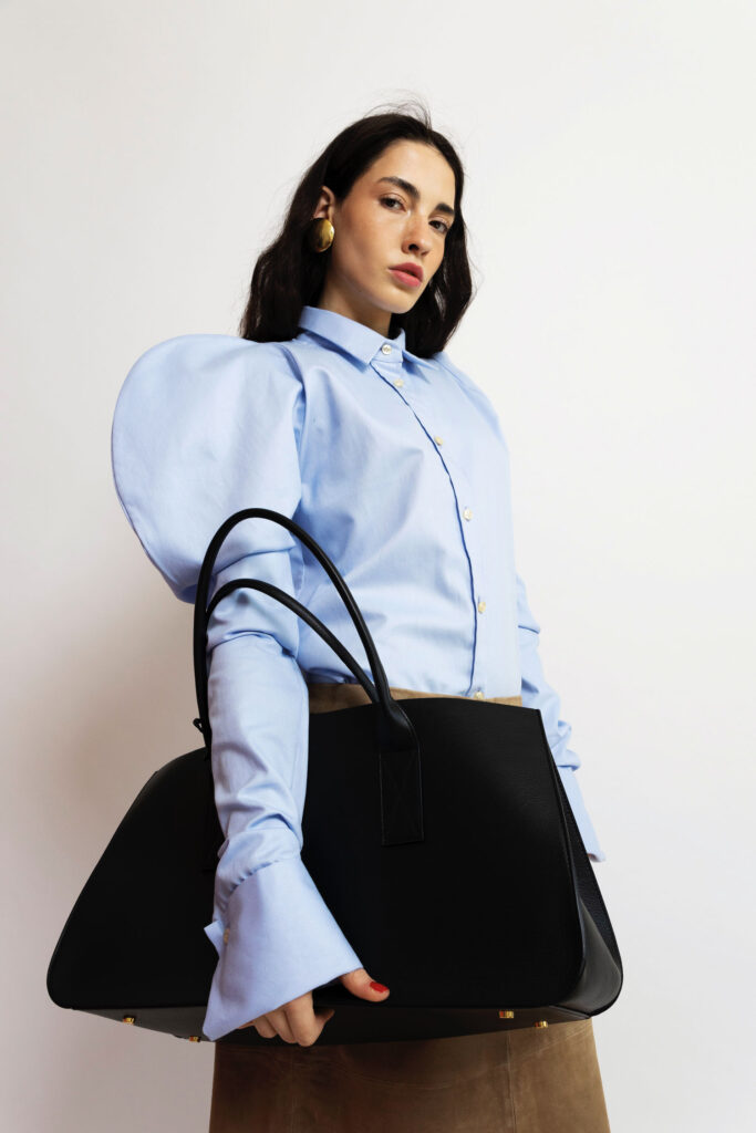 Julia Skergeth Handtaschen: Elegante, große, schwarze Tasche präsentiert von einer dunkelhaarigen Frau mit weitem, hellblauen Hemd.