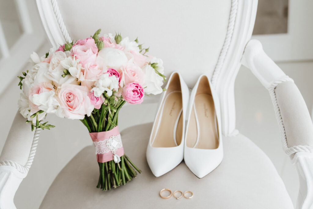 Brautstrauß mit weißen und rosa Blumen liegt auf einem antiken Stuhl mit Ringen und weißen Stöckelschuhen.