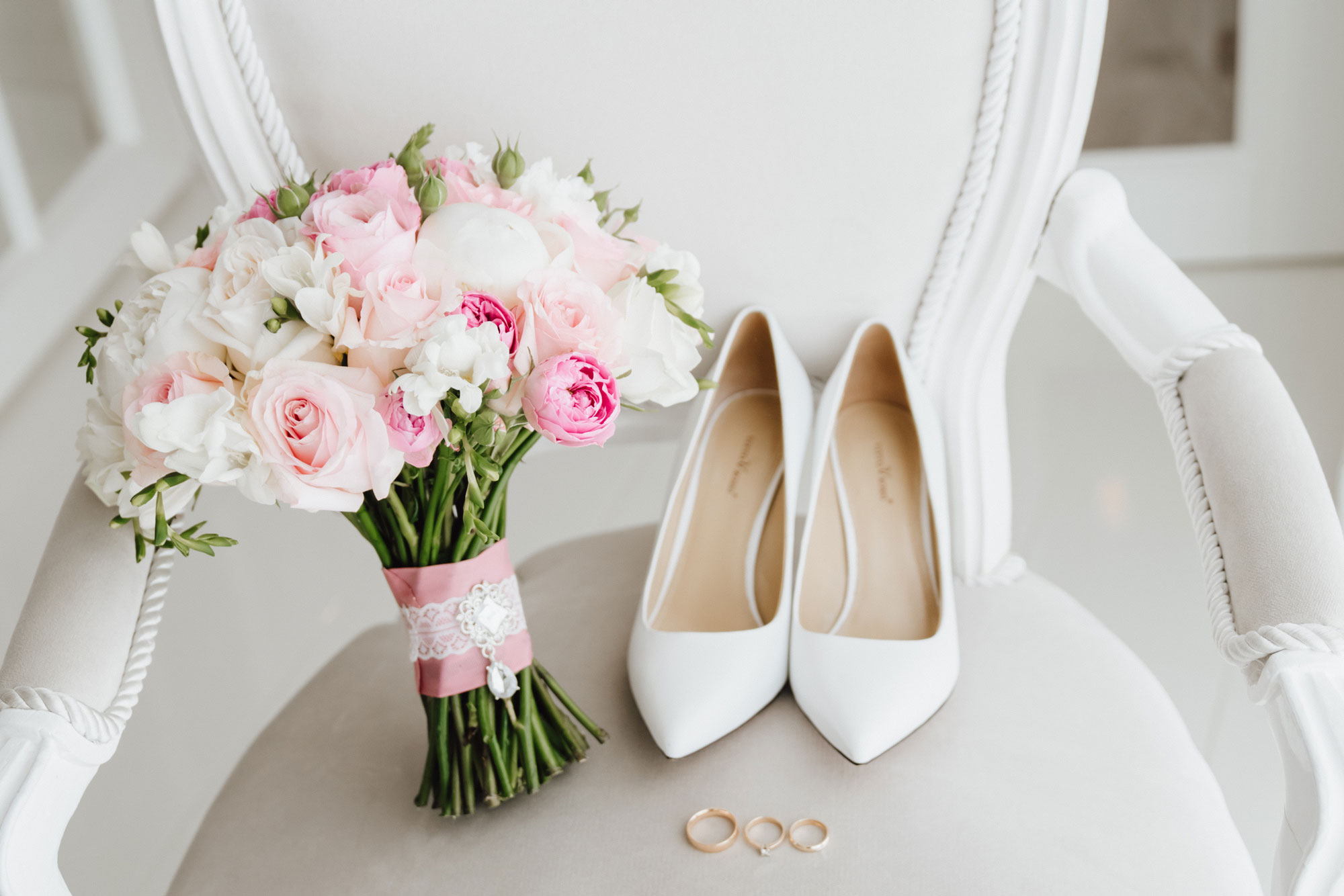 Brautstrauß mit weißen und rosa Blumen liegt auf einem antiken Stuhl mit Ringen und weißen Stöckelschuhen.