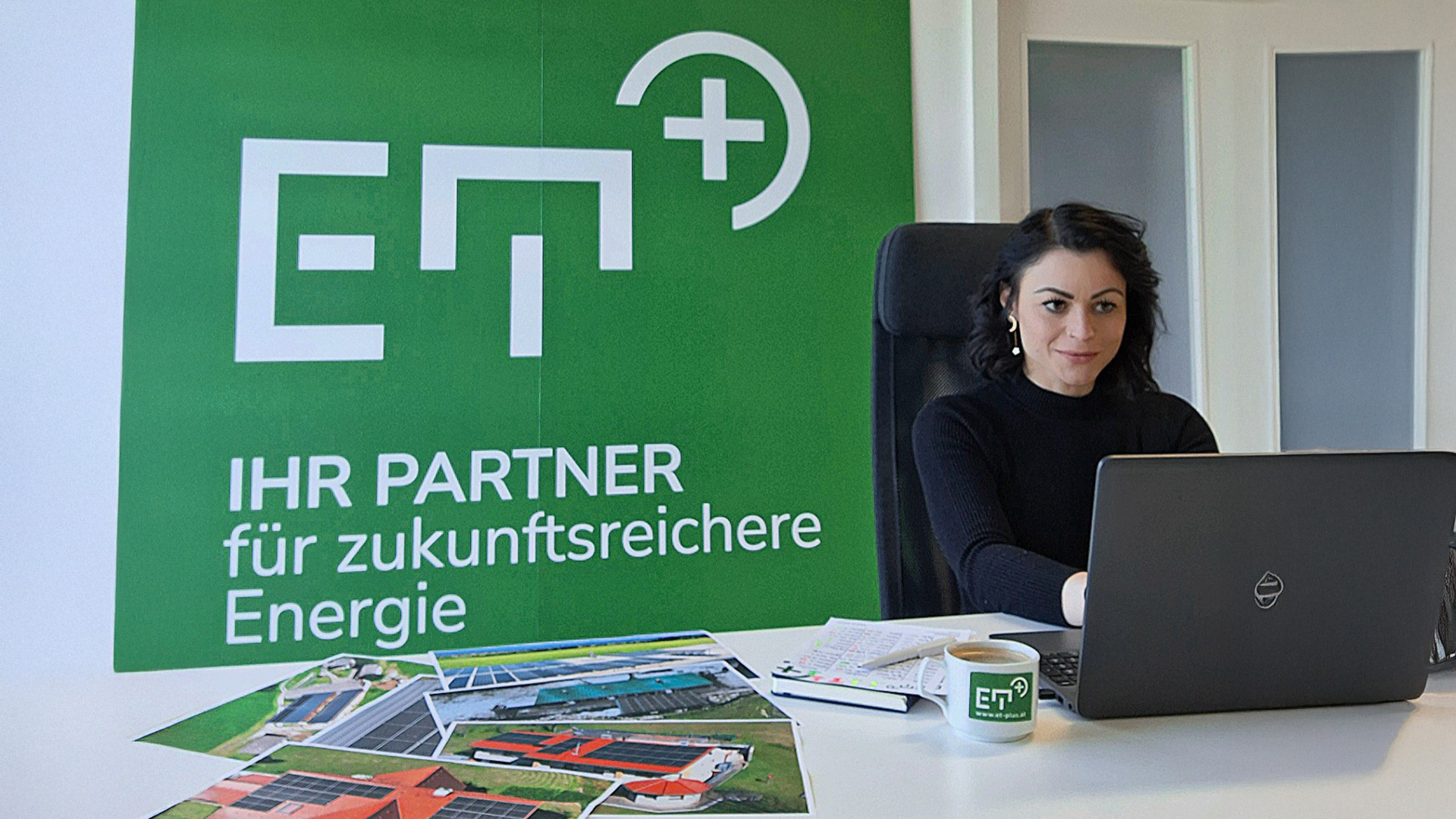 Frau arbeitet an einem Laptop mit Bildern neben sich am Tisch liegen und einem großen grünen Plakat an der Wand mit der Aufschrift "ET PLUS – IHR PARTNER für zukunftsreichere Energie".