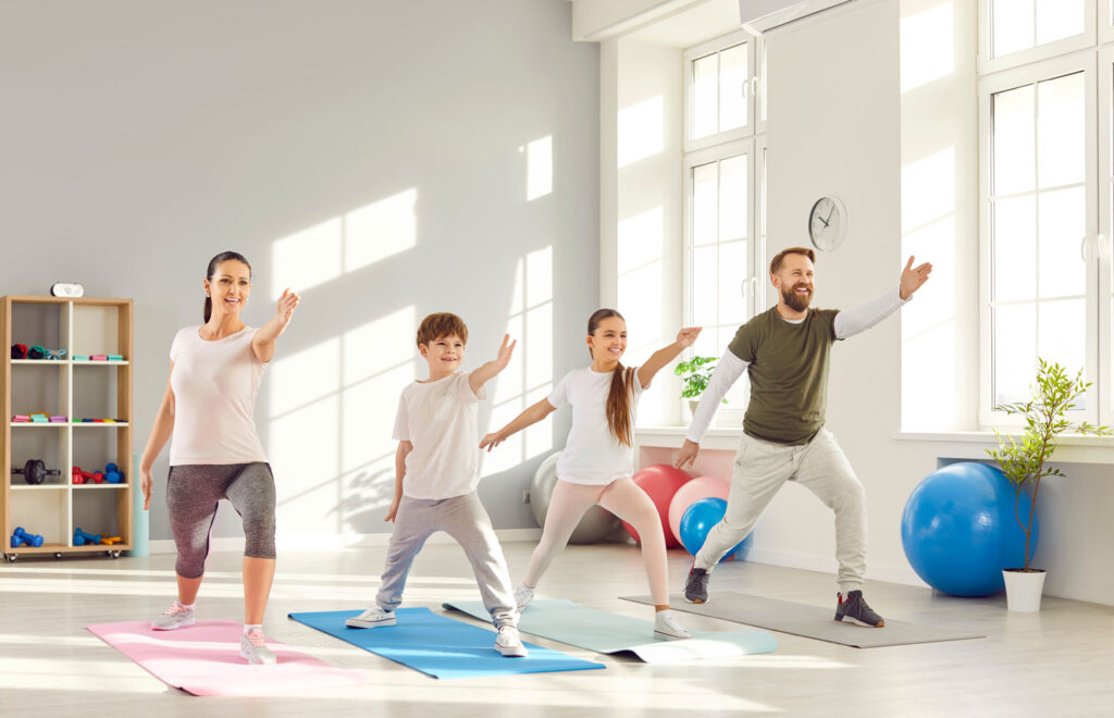 Zwei erwachsene und zwei Kinder machen sportliche Übungen in einem hellen Turnraum.