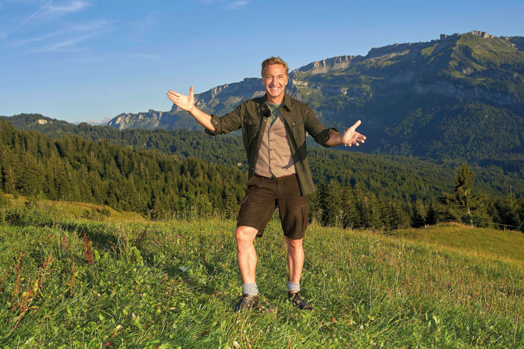 Hans Knauß in steirischer Tracht auf einer Weide mit Bergen im Hintergrund.
