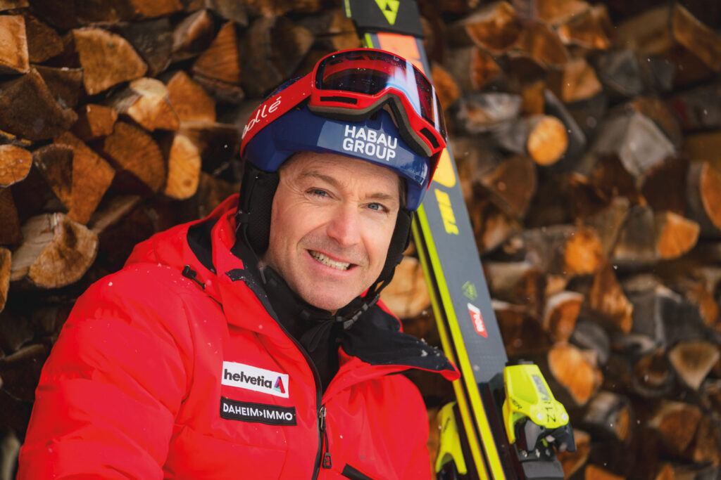 Hans in roter Ski-Montur miit Skiern in der Hand und Holzklötzen im Hintergrund.