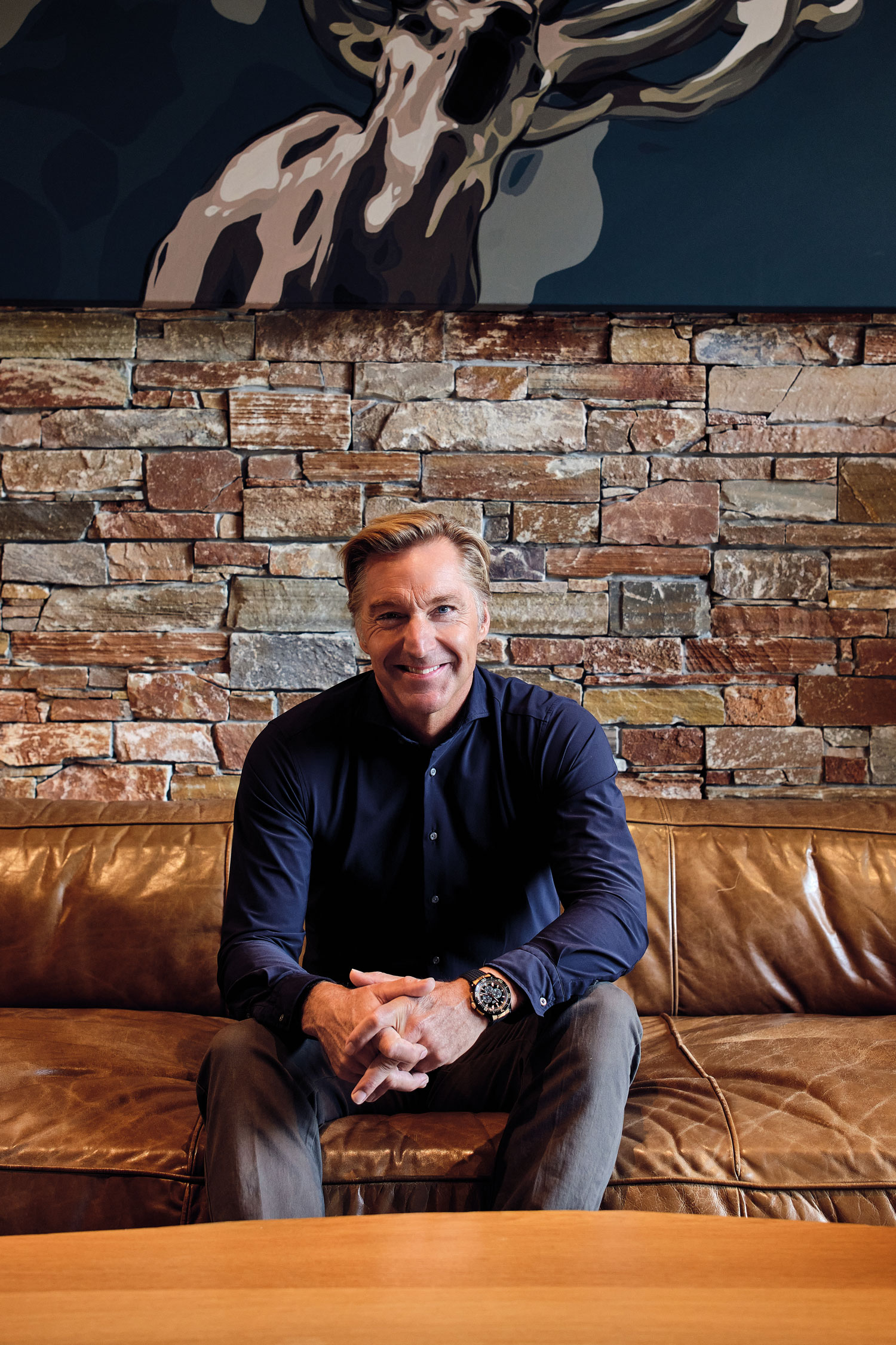 Hans sitzt lachend auf einer ledernen Couch mit Backstein-Wand im Hintergrund.