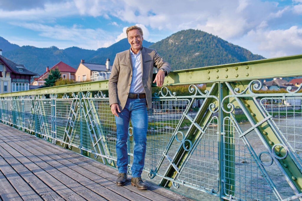 Hans Knauß auf einer grün-türkisen Brücke mit Schnörkeln und Dorf sowie Bergen im Hintergrund.