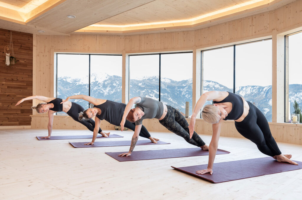 Vier Damen machen in einem hölzernen Raum vor verschneiter, bergiger Aussicht Yoga-Übungen.