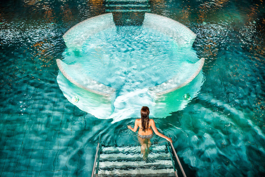 Ansicht von oben auf türkises Wasser in einem Thermalbecken und eine Frau, die über Treppen hineingeht.