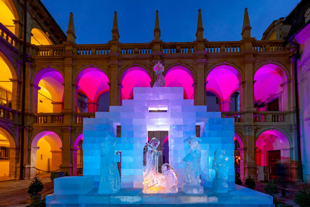 Advent in Graz: Die blau beleuchtete Eiskrippe im Innenhof mit pink-oranger Beleuchtung in den Arkaden im Hintergrund.