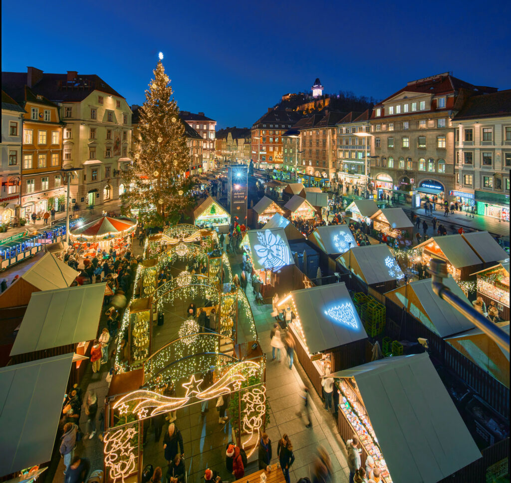 Advent in Graz: Der Grazer Hauptplatz mit weihnachtlich beleuchtetem Christkindlmarkt und Schlossberg mit Uhrturm im Hintergrund.
