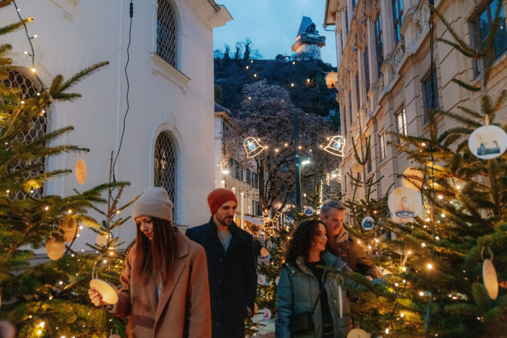 Eine Grazer Gasse mit beleuchteten Christbäumen und Menschen, im Hintergrund der Schlossberg mit Uhrturm.