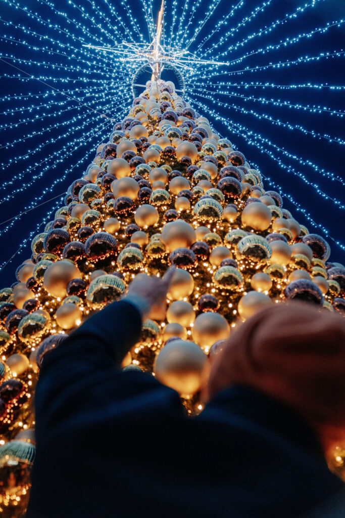 Eine unscharfe Person im Vordergrund zeigt auf einen hohen Christbaum aus gold-braunen Christbaumkugeln mit weißer Lichterketten-Beleuchtung am Spitz.