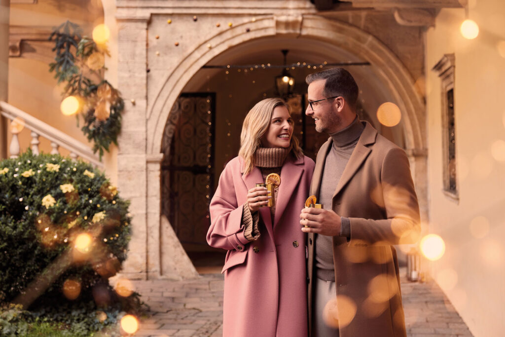Ausflugsziele Steiermark: Winterlich gekleidetes Pärchen steht fröhlich mit Punsch in einem weihnachtlichen Innenhof.