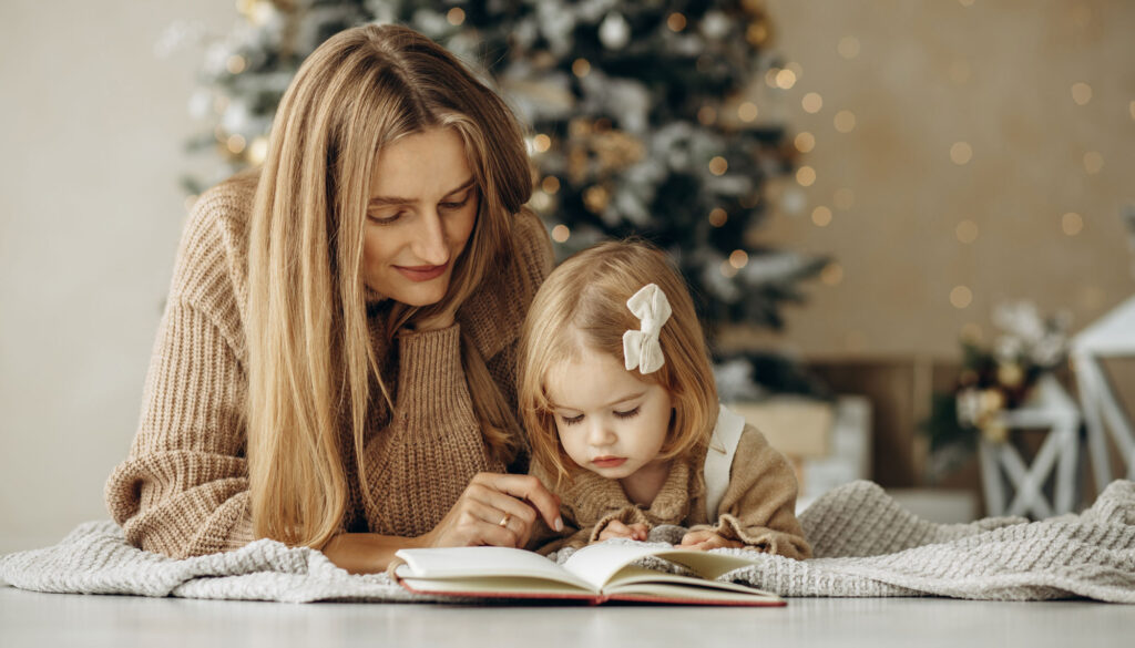 Weihnachtsbücher: Mutter liest mit Tochter auf einer Decke ein Buch mit einem Christbaum im Hintergrund.