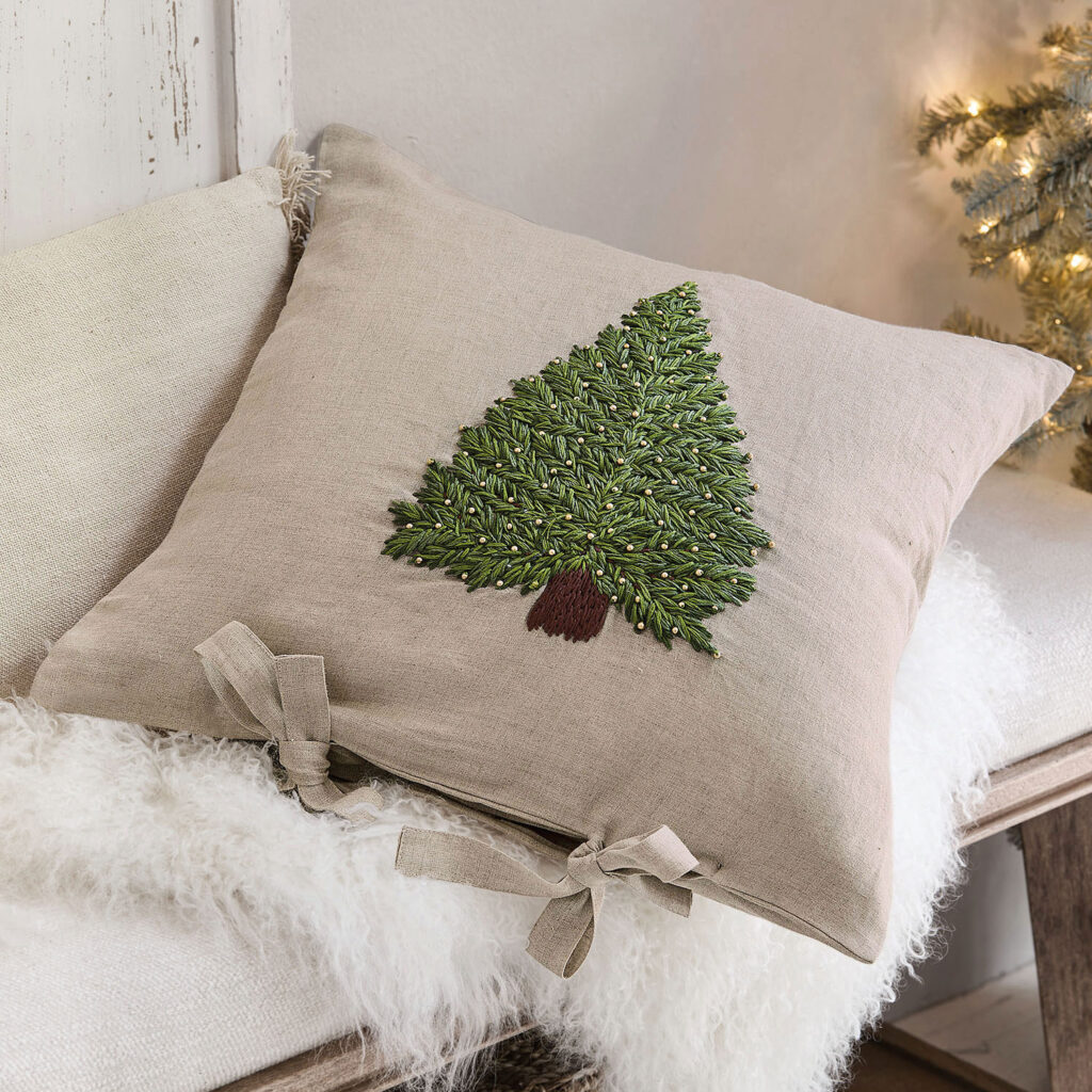 Weihnachtsdeko: Ein beiges Kissen mit gesticktem Tannenbaun lieg auf einem Sofa mit leuchtendem Christbaum im Hintergrund.