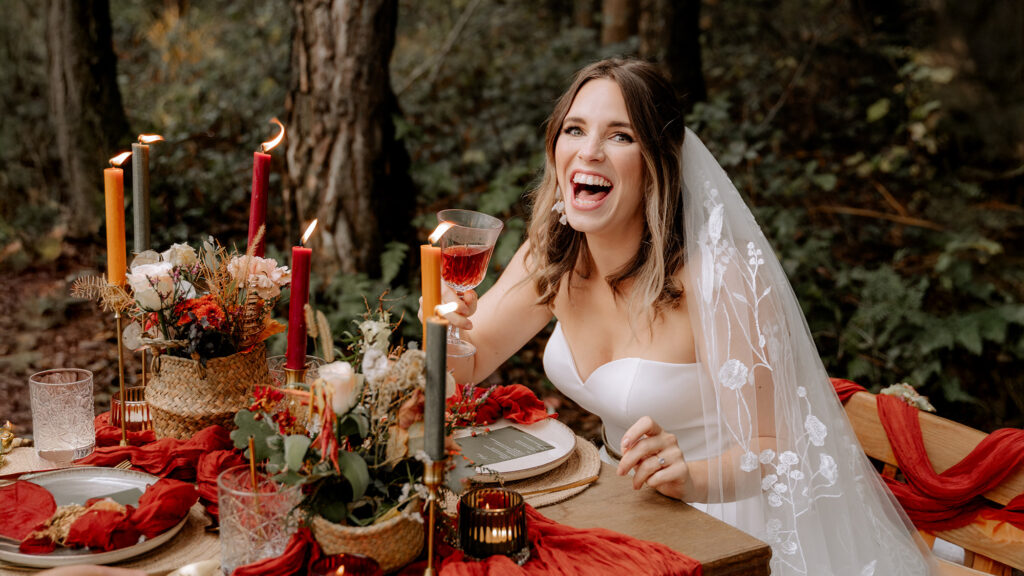 Kerstin Pomper von Kamerafräulein fängt die einzigartige Stimmung von Herbsthochzeiten ein für authentische Hochzeitsfotos: Hier sitzt eine Braut lachend an einem rot eingedeckten Tisch im Wald.