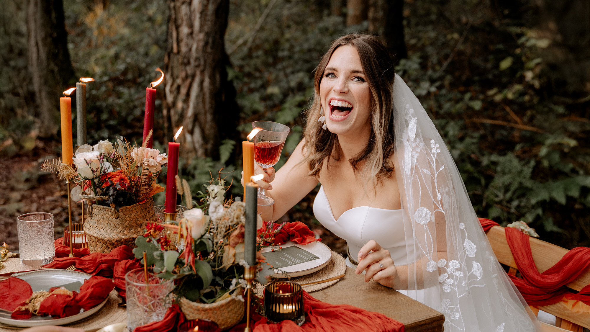 Kerstin Pomper von Kamerafräulein fängt die einzigartige Stimmung von Herbsthochzeiten ein für authentische Hochzeitsfotos: Hier sitzt eine Braut lachend an einem rot eingedeckten Tisch im Wald.