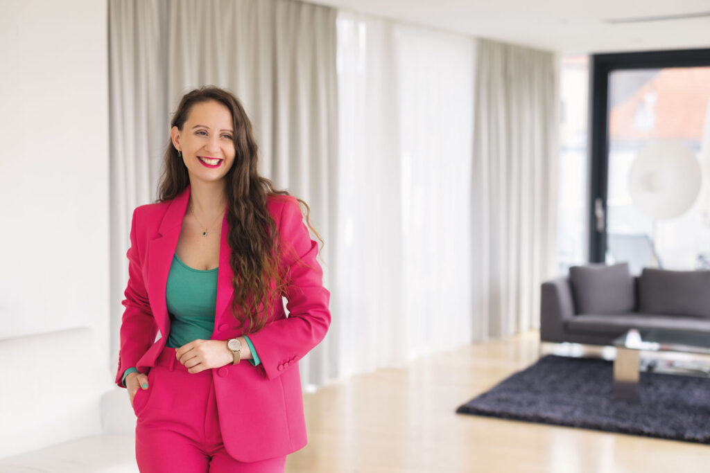 Portrait von Sara Obereder mit pinkem Anzug und türkisem Shirt in einem Wohnzimmer.