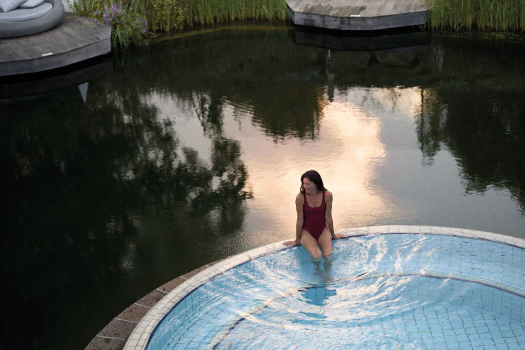 Eine Frau sitzt im roten Badeanzug am Rande eines Pools, das in einem Teich ist.