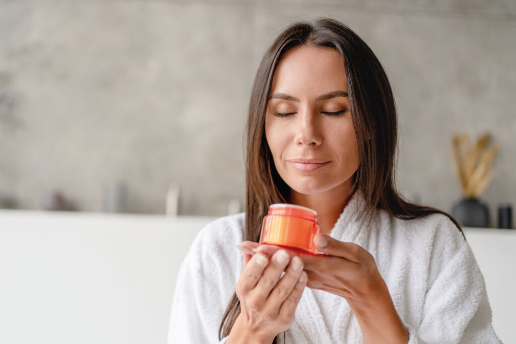 Frau in Bademantel riecht mit geschlossenen Augen an einem orangen Döschen.