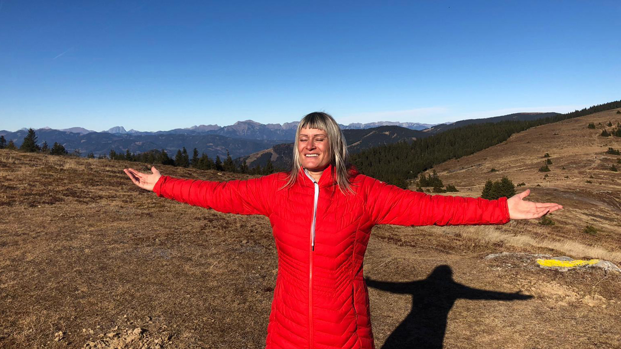 Heide Pötz trägt eine rote Jacke und streckt die Arme aus mit Bergen im Hintergrund.
