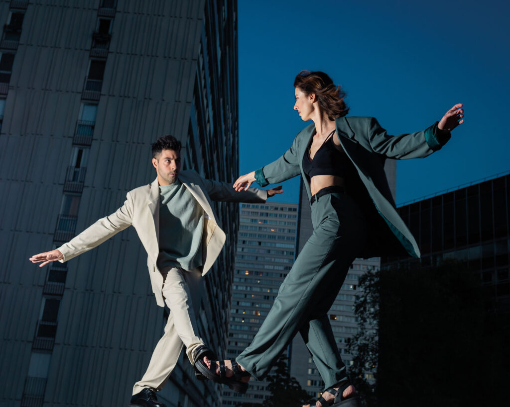 Ein Mann und eine Frau tanzen in Anzügen im Freien mit Hochhäusern im Hintergrund.