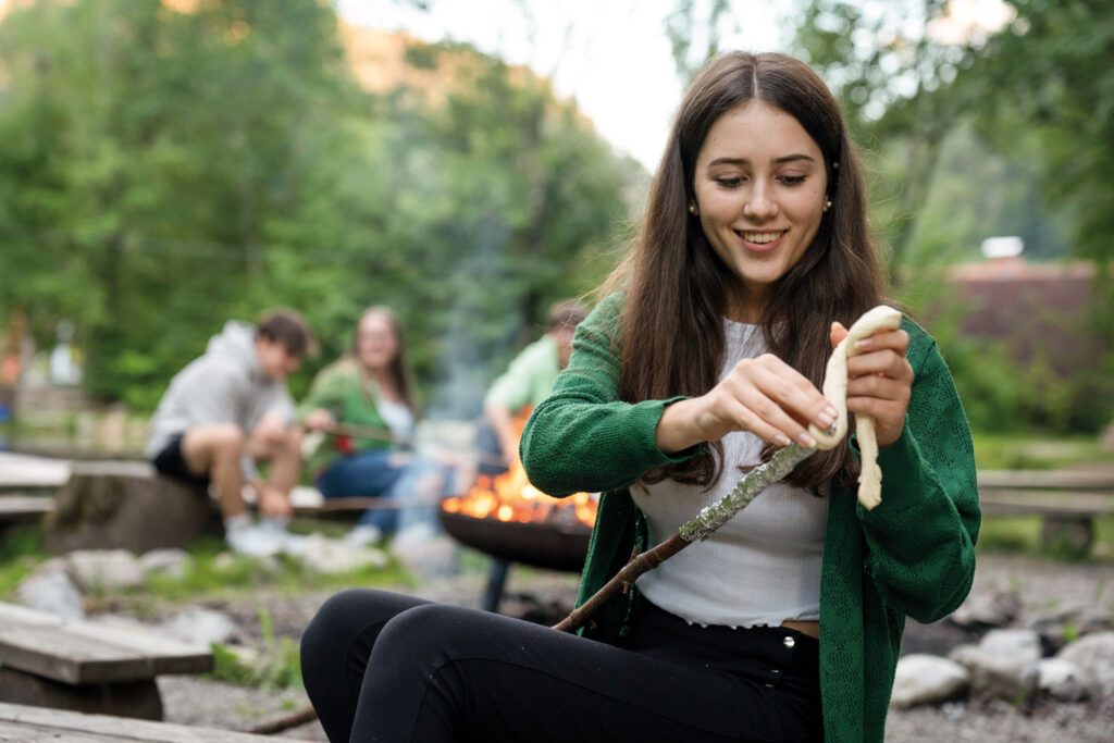 Eine junge Frau bringt an einem Lagerfeuer Stockbrot auf einem Stock an.