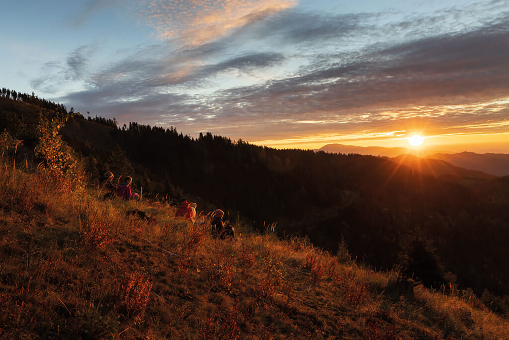 Menschen sitzen auf einer Wiese am Berg und beobachten den Sonnenuntergang.