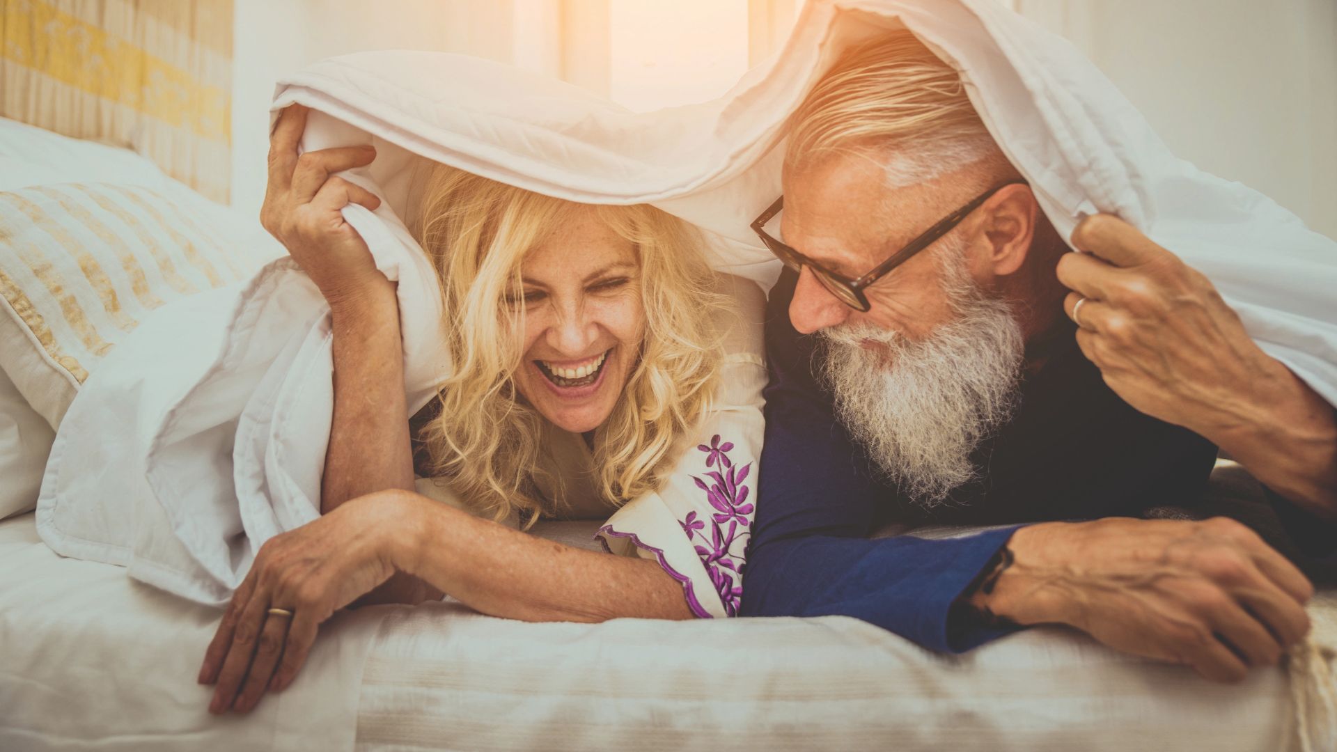 Älteres Paar liegt lachend im Bett unter der Decke