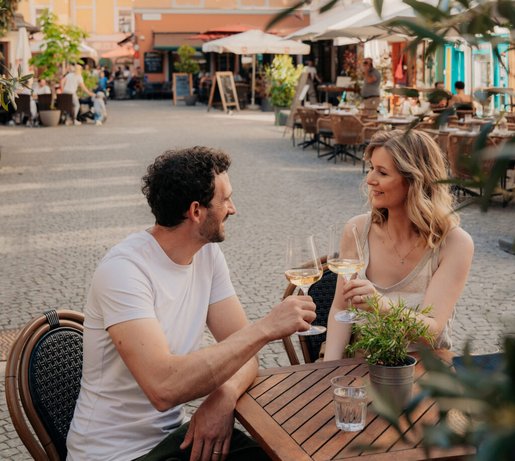 Ein Mann und eine Frau sitzen in einer gemützlichen Gasse mit einem Gläschen Wein.