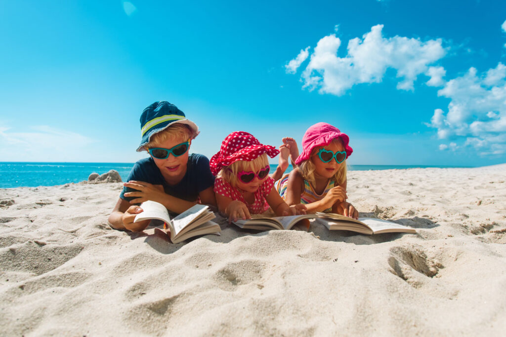 Ferienbücher: Drei Kinder lesen Bücher am Strand.